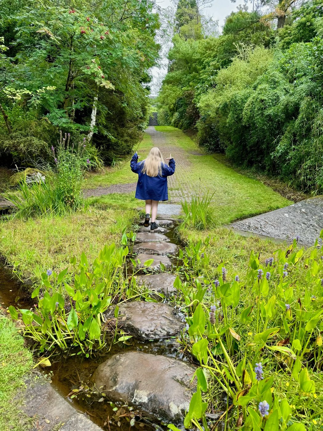 A figure crossing stepping stones over a stream in a lush green garden
